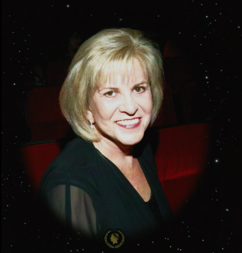 A portrait of author Amelia Sterling smiling at the camera. She has shoulder-length blonde hair and is wearing a black top with sheer sleeves and small drop earrings. The background is dark, showing blurred red theater seats, and is framed by a black vignette with a starry effect.