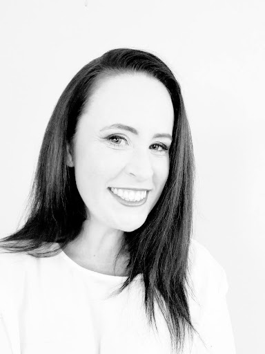 A black and white headshot of author Megan Lane smiling brightly at the camera. She has long, dark straight hair and is wearing a simple light-colored top against a plain white background.