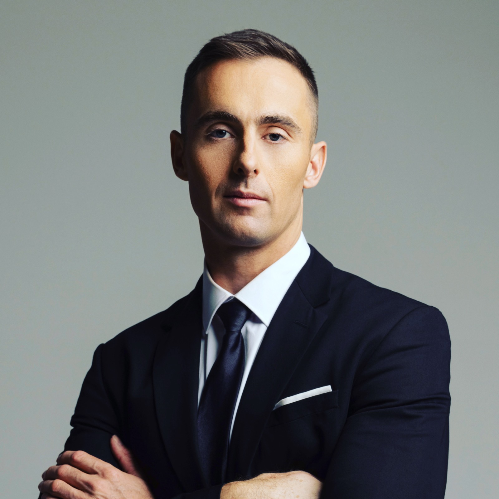 A professional studio portrait of a man, identified as Spencer Tarring, against a solid gray background. He is wearing a dark, tailored suit, a white dress shirt, and a dark tie with a subtle sheen. He stands with his arms crossed and looks directly into the camera with a neutral, confident expression.
