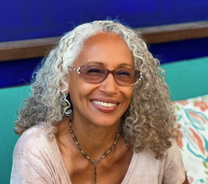 A professional portrait of author Carine Fabius smiling warmly. She has long, curly grey hair, wears glasses with decorative frames, and a beaded necklace. She is seated against a vibrant blue background with a floral patterned pillow visible behind her.
