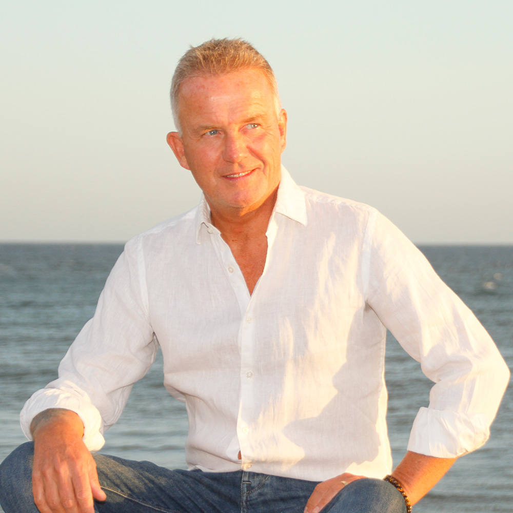 A professional photograph of Glenn Harrold. He is wearing a white button-down shirt and jeans, sitting outdoors in front of a calm ocean at sunset.