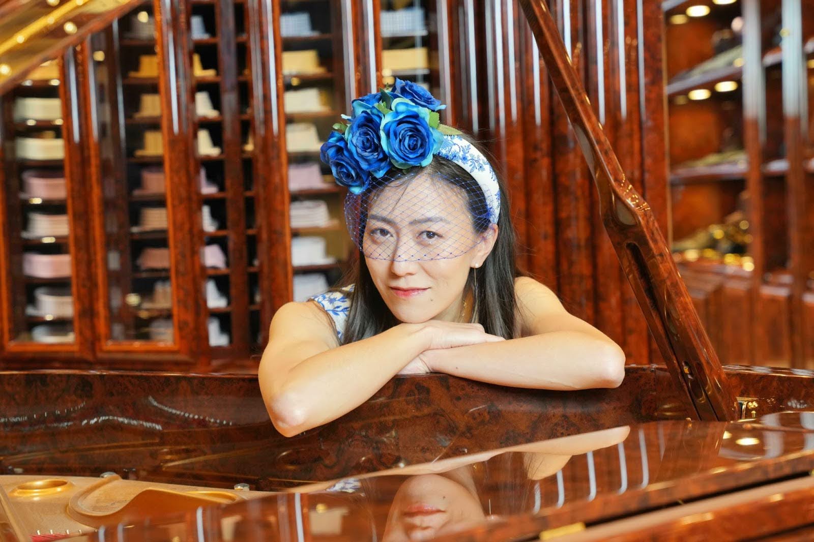 Portrait of Miist, an Asian woman with long dark hair wearing a white off-shoulder top and a headpiece of large blue roses on a patterned band, smiling softly with mesh veil over her face, leaning forward with arms crossed on a polished wooden grand piano in a luxurious wood-paneled room lined with shelves.